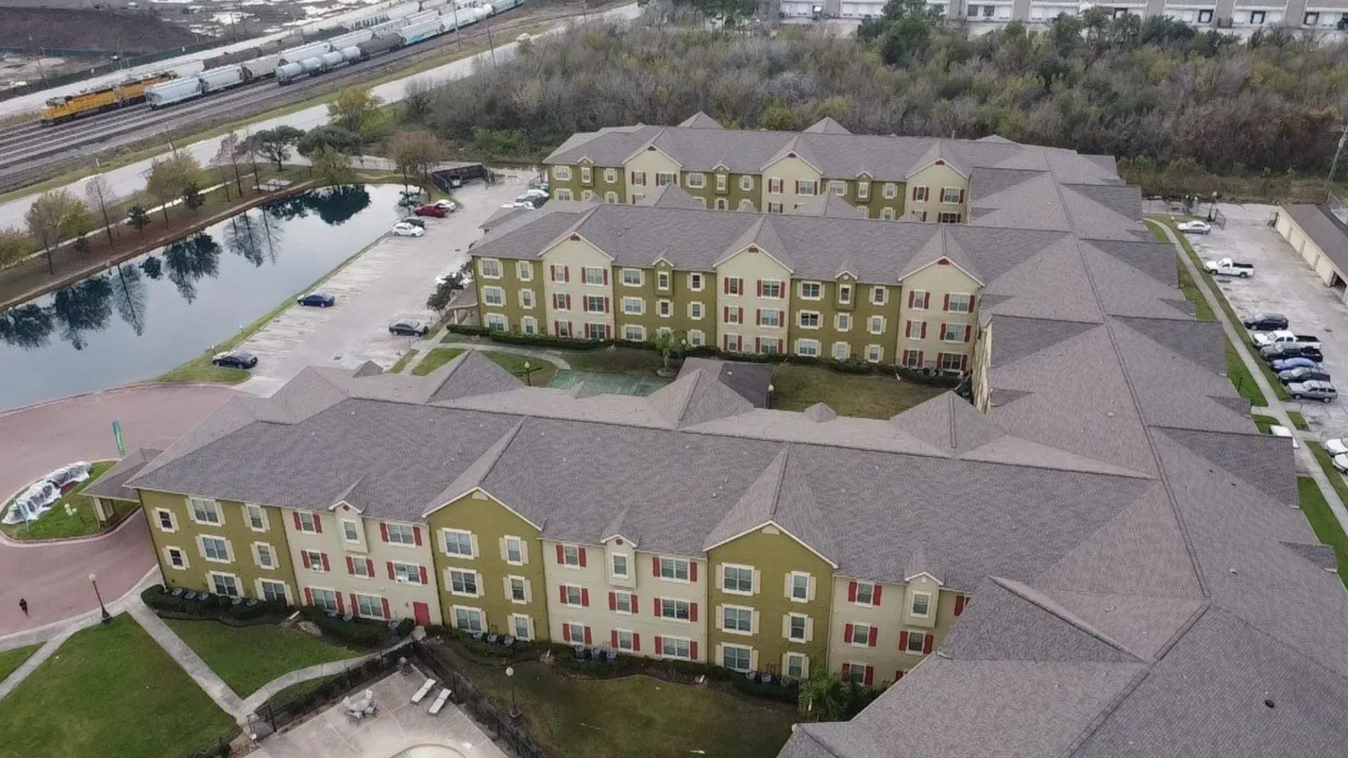 A large, traditional, 3-story, olive green and cream garden-style apartment complex with red shutters and connected buildings covered with GAF HDZ shingle roofs off a Highway in Houston, TX.