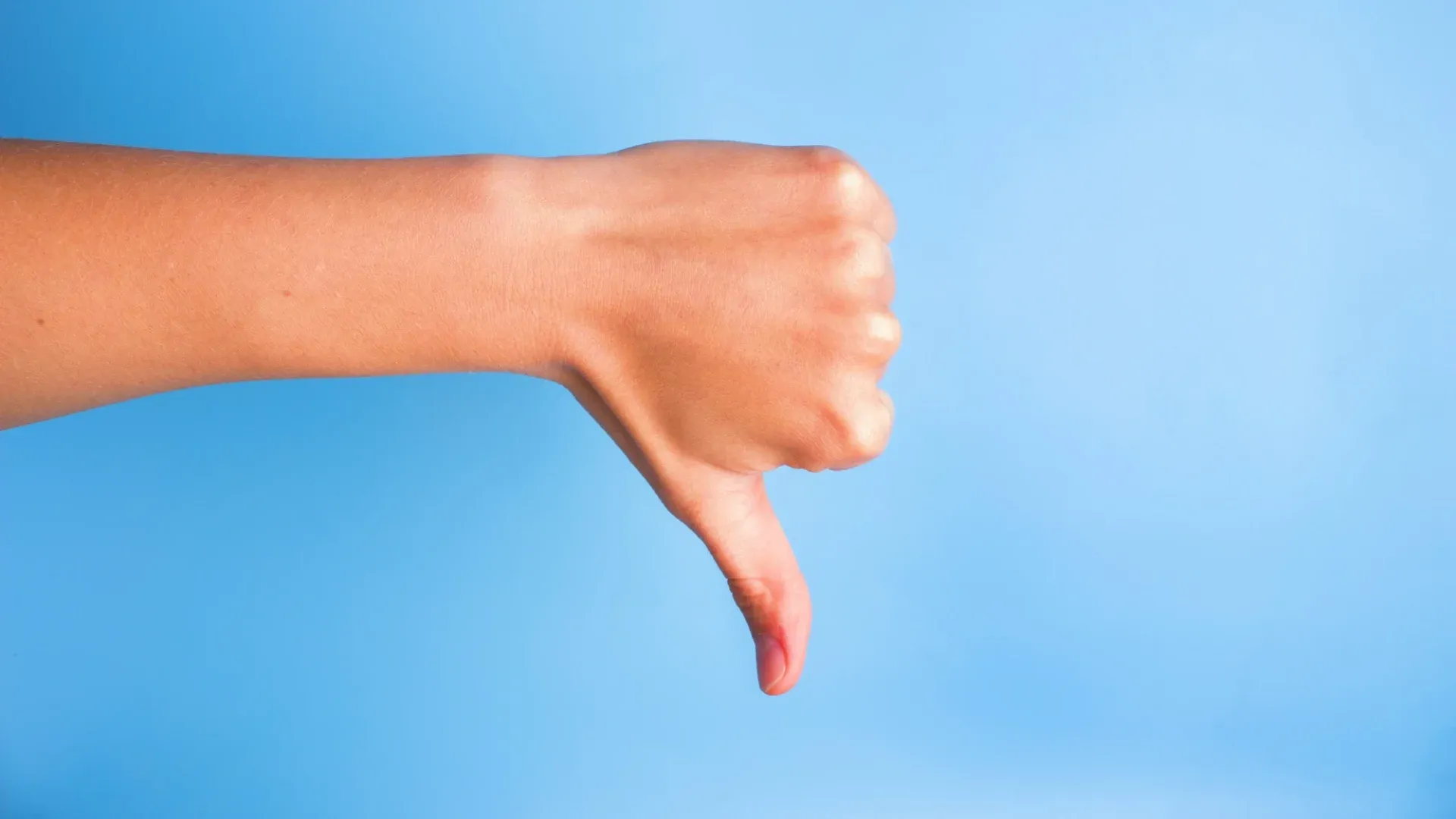 A closeup of a woman’s hand gesturing a thumbs down against a sky-blue background