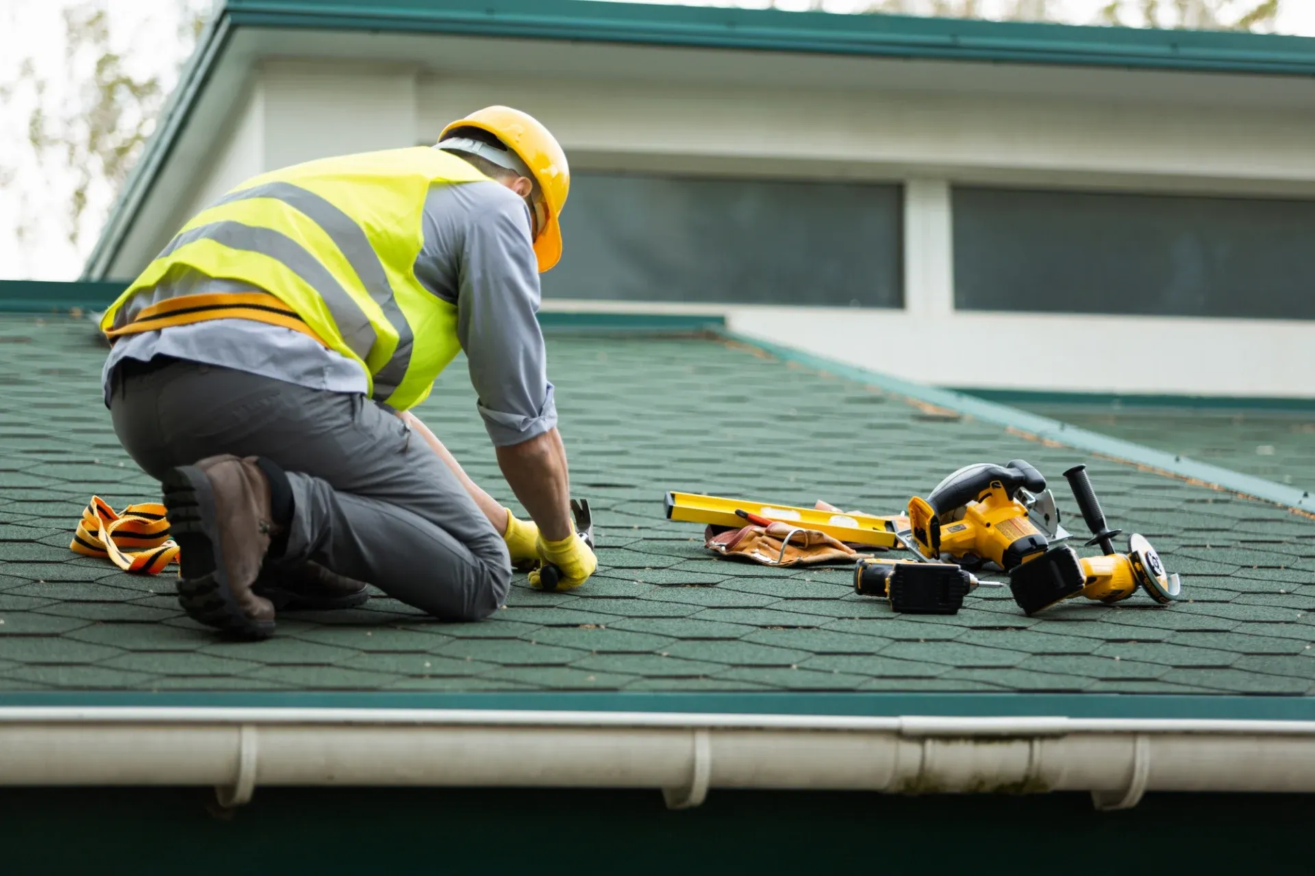 Roofer in safety gear repairing green asphalt shingle roof
